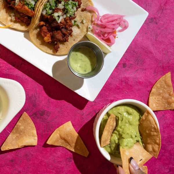 Friducha's Mexican tacos with guacamole and chips on a vibrant pink background.