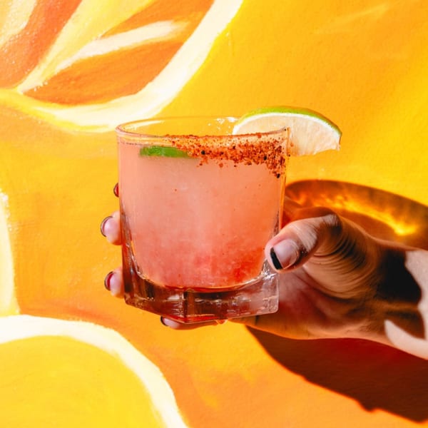 Hand holding a Friducha pink margarita with a chili-lime rim and lime wedge against a vibrant orange wall.