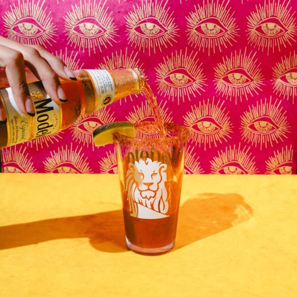 A hand pours a Modelo beer into a Friducha glass with a chili-lime rim and lime wedge.