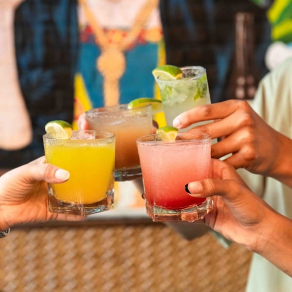 Four hands holding colorful Friducha cocktails with lime garnishes, clinking glasses in a toast.