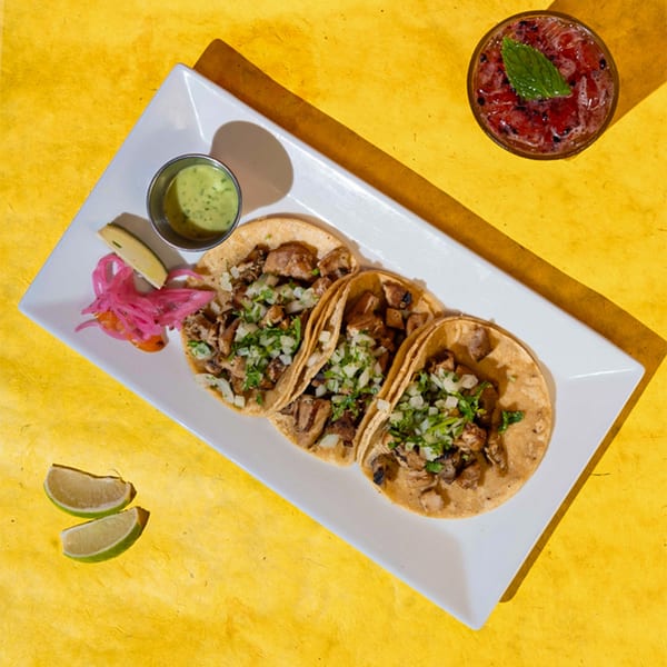 Three Friducha tacos with meat, onions, and cilantro, with a side of green sauce and pickled onions.