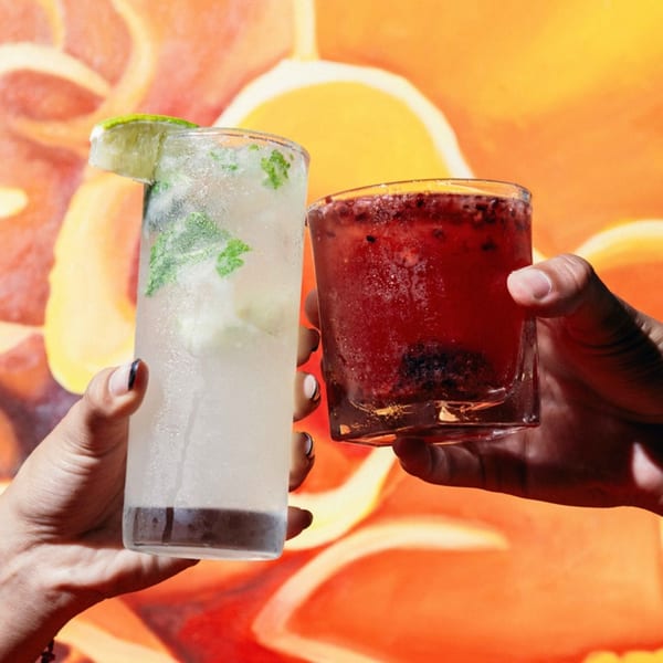 Two hands holding a mojito and a red berry cocktail against an orange background.