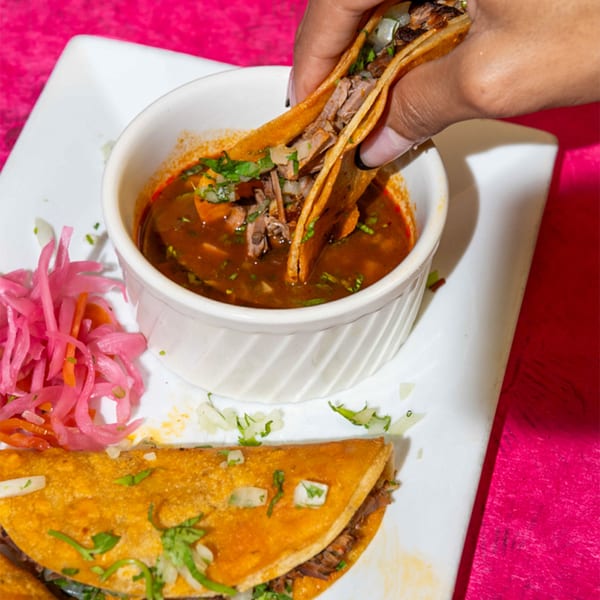 Hand dipping a Friducha birria taco into a bowl of consome, with pickled onions on the side.