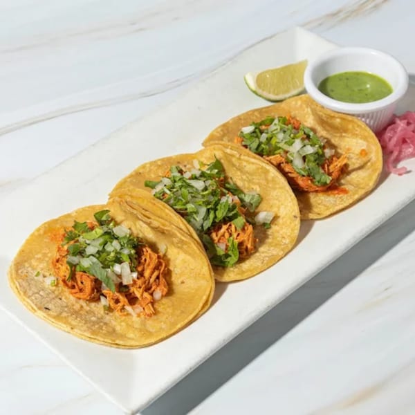 Three Friducha Mexican tacos with shredded chicken, cilantro, and onions, served with lime and green sauce.